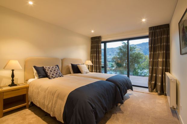 A warm, contemporary bedroom with two single beds dressed in cream and dark navy bedding, matching bedside tables and lamps, beige carpet, and floor-to-ceiling sliding glass doors opening to a scenic mountain and wooded view. Neutral walls, full-height curtains, and simple framed artwork create a comfortable, refined interior.