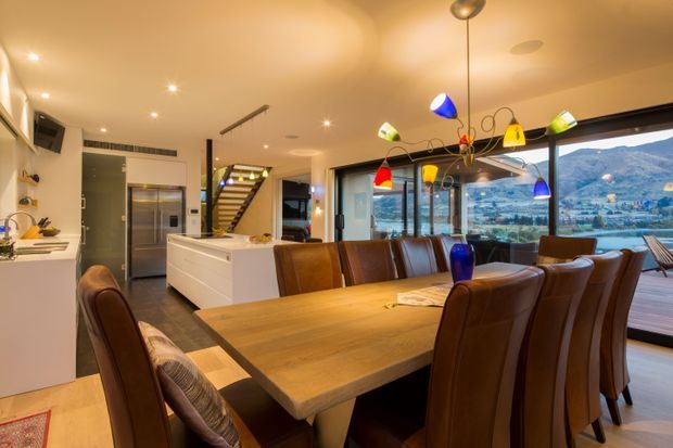 Open-plan contemporary dining and kitchen area with a large timber dining table surrounded by upholstered leather dining chairs. A white island kitchen, stainless-steel refrigerator, stairway, extensive glazing and a colourful multi-light pendant are visible. The windows overlook a mountainous waterfront landscape.