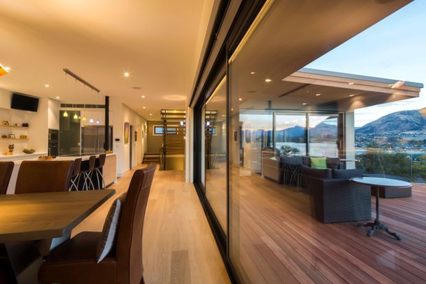 A contemporary open-plan residential interior opening onto a spacious timber deck through full-height sliding glass doors. The image shows a dining area with upholstered chairs, a kitchen with island seating and pendant lighting, and an outdoor lounge overlooking a mountainous landscape and water. Warm timber flooring and decking contrast with dark-framed glazing and light walls.