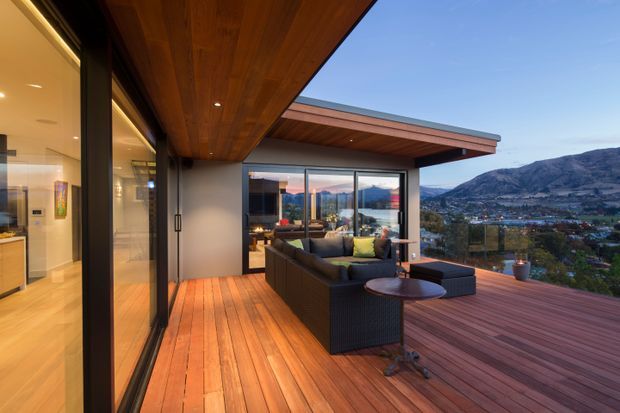 A modern covered outdoor terrace overlooking a dramatic mountain and lakeside landscape. The space features warm timber soffits and decking, expansive black-framed glazing, and a dark outdoor sectional lounge with a low round table. The flat-roofed structure creates a sheltered, open-air living area with strong indoor-outdoor connection.