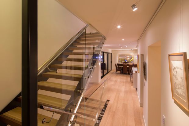 A warm contemporary interior hallway with pale timber flooring, a floating open-riser staircase, and clear glass balustrades with dark metal framing. The corridor leads toward a dining area, with recessed and track lighting, framed artwork, and a partial view of adjoining rooms.