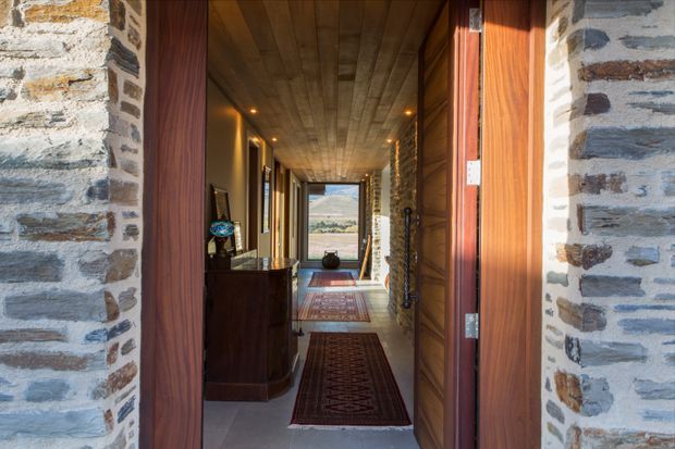 View through an open timber entry door into a long, narrow hallway. The hallway features exposed natural stone walls, warm timber wall and ceiling lining, recessed downlights, timber joinery, patterned runners, and a glazed door or window at the far end with a landscape view beyond.
