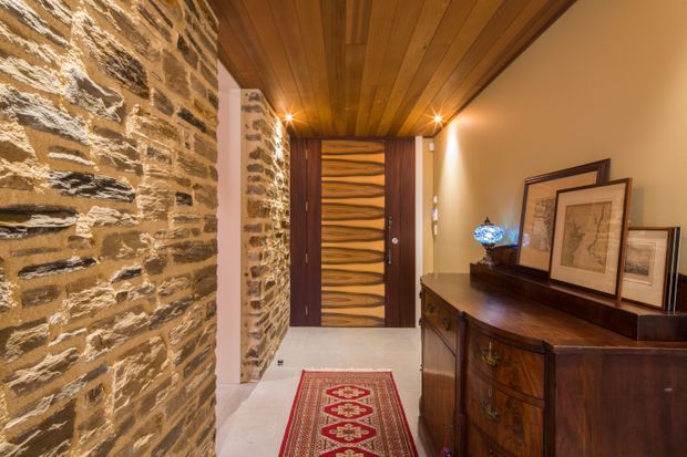 A warm, richly detailed entry hall with a rugged exposed stone feature wall, timber-plank ceiling, pale flooring, and a striking dark timber front door with a wavy inset design. A traditional dark wood sideboard, framed artwork, decorative lamp, and patterned runner add character and heritage-inspired warmth.