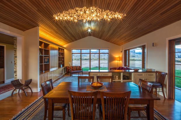 Warm open-plan dining and living space with a large timber dining table and slatted-back chairs, extensive built-in timber shelving, a dramatic faceted crystal chandelier, and a richly detailed timber-lined vaulted ceiling. Large glazed doors and windows connect the room to the landscape outside.