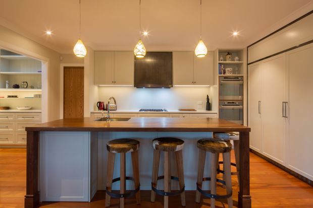 Warm, traditional-style kitchen with a large central island and three timber bar stools. The room features white built-in cabinetry, a dark rangehood, integrated appliances, open shelving, timber flooring, pendant lights, and a timber island benchtop. A pantry or secondary kitchen area is visible through the opening on the left.