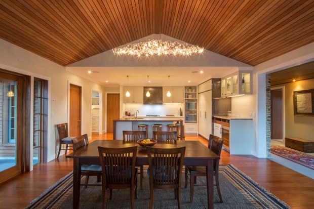 Warm open-plan dining and kitchen interior with a large dark timber dining table and chairs in the foreground, a central kitchen island, extensive white cabinetry, timber flooring, and a dramatic slatted timber ceiling forming a vaulted geometric canopy. Recessed lighting and pendant lights provide a welcoming glow, while a wide opening connects to an adjacent living area.