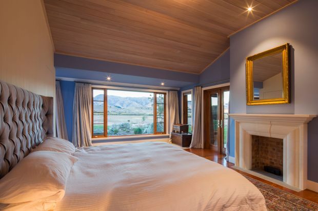 A warm, traditional bedroom with a large upholstered bed, deep blue walls, timber-lined ceiling, timber-framed windows, and a white classical fireplace surround. The room opens to expansive mountain views through large windows and glazed doors, with draped curtains, a gold-framed mirror, and layered patterned flooring adding a refined, luxurious character.