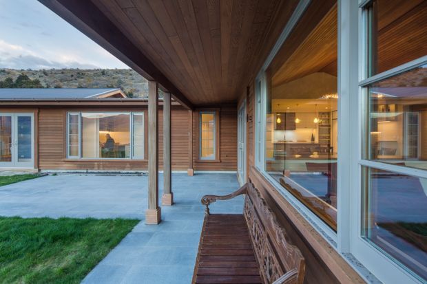 A timber-clad residential courtyard viewed from a covered outdoor walkway or veranda. The scene features warm timber soffits and walls, large white-framed windows, a built-in or fixed timber bench, concrete paving, and a small grass courtyard. Interior kitchen and living areas are visible through the glazing, while a low mountain or rocky landscape appears in the distance.