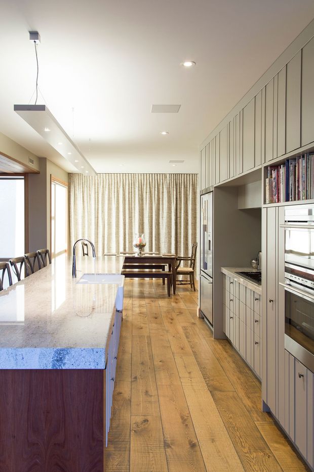 Bright, elongated open-plan kitchen and dining area with warm timber flooring, a large pale stone island, dark timber island cabinetry, and extensive light grey built-in cabinetry along the right wall. Integrated oven, cooktop, shelving, and pantry-style storage are visible, with a dining table set in front of full-height patterned curtains. Natural light enters through tall windows on the left, complemented by recessed ceiling lights and a linear pendant.