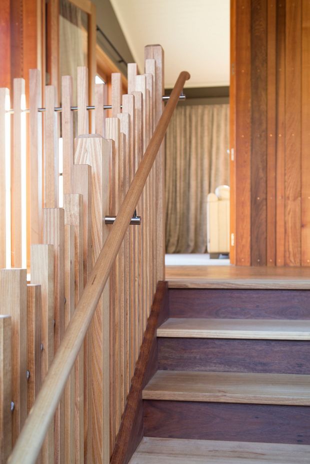 Close-up interior view of a timber staircase with richly stained dark timber treads and risers, lighter timber stair nosings, a vertical timber balustrade, and a matching rounded timber handrail. The stairs lead toward a warmly finished landing and corridor with timber wall panelling and a pale ceiling.