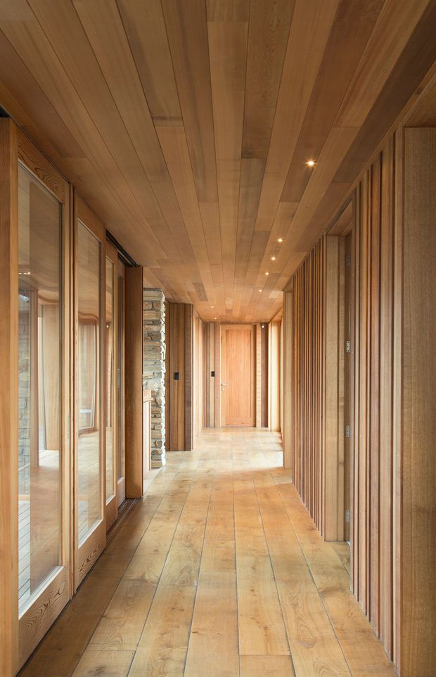 A long, warm-toned interior hallway with extensive timber lining on the ceiling, floor, doors and wall panels. Full-height glazed openings with timber frames line the left side, while vertical timber battens create a textured wall on the right. A natural stacked-stone feature wall is visible midway along the corridor, with recessed downlights leading toward a timber door at the end.