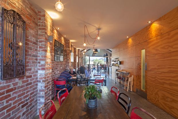 A long, narrow commercial dining space or café with exposed red brick walls, black wrought-iron detailing, warm timber wall panelling, polished concrete flooring, and rows of small tables with colourful metal chairs. Pendant and recessed lighting create a warm industrial atmosphere, with a service or bar area visible toward the rear.