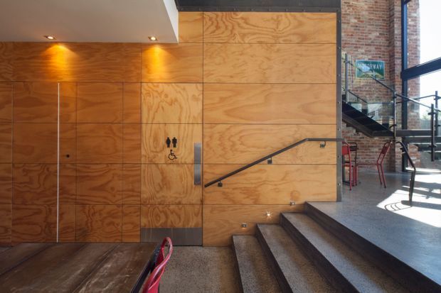 Interior view of an industrial-style commercial space with a plywood-clad wall and integrated restroom doors, exposed red-brick walls, dark metal handrails, concrete steps, and a raised seating or circulation area. A partially visible café-style table and chairs appear on the upper level, while recessed ceiling lights illuminate the timber wall.