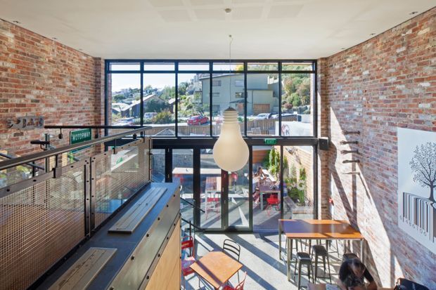 A bright, multi-level commercial interior with exposed red brick walls, black-framed floor-to-ceiling glazing, and an industrial-style mezzanine overlooking a lower seating area. Metal mesh balustrades, timber-topped bench seating, café tables and chairs, and a large pendant light define the space. Urban buildings are visible through the glazing.