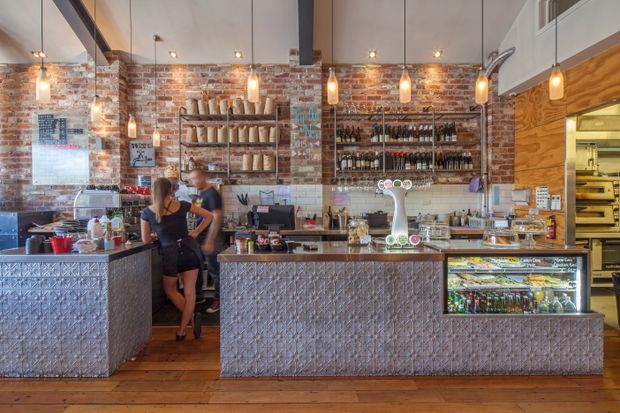 A warmly lit café or bar interior with exposed red brick walls, timber flooring, pendant lights, and a long patterned tiled service counter. Shelving displays bottles and bread behind the counter, with refrigerated food and drink cases in the foreground and a commercial kitchen visible at right.