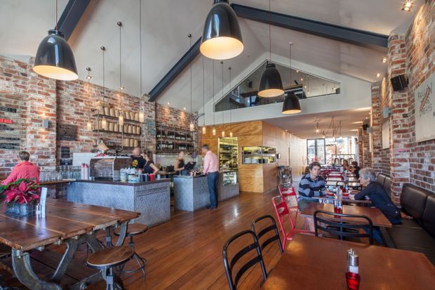 A spacious industrial-style café or restaurant interior with exposed red brick walls, dark steel beams, suspended black pendant lights, timber flooring, communal timber tables, and a central stone-finished service counter. Shelving displays bottles and café supplies, while a mezzanine-like opening and large front glazing add openness and daylight.
