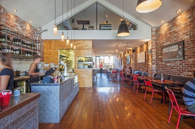 A lively industrial-style café or restaurant interior with exposed red brick walls, polished timber plank flooring, a long bar with textured metal cladding, open shelving stocked with bottles, pendant lighting, and red dining chairs along the right wall. The space features a high pitched ceiling with a glazed mezzanine or upper-level opening, creating a spacious hospitality setting.