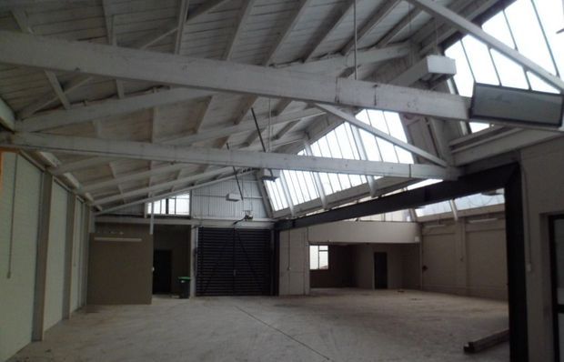A large vacant industrial-style interior with a broad concrete floor, exposed white-painted timber roof trusses and beams, corrugated wall panels, high clerestory glazing, and multiple large openings or mezzanine-like structures. The space appears to be an unfinished warehouse or converted commercial building with abundant overhead daylight.