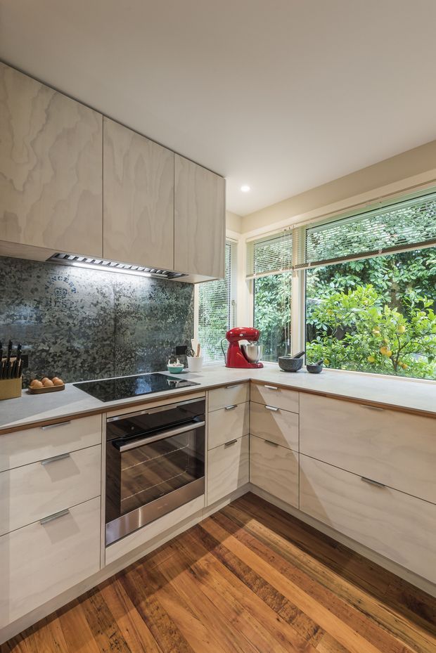 A compact L-shaped kitchen with light timber cabinetry, a pale benchtop, dark mottled backsplash, integrated stainless-steel oven and black induction cooktop. Large corner windows with horizontal blinds overlook dense greenery, while warm timber flooring and a red stand mixer add contrast.