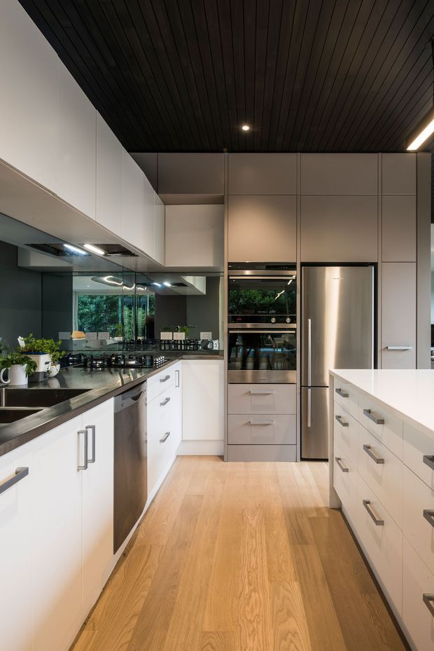A contemporary galley-style kitchen with extensive white flat-panel cabinetry, integrated stainless-steel appliances, dark stone benchtops, a reflective splashback, and warm timber flooring. The ceiling is clad in dark timber battens with recessed and linear lighting. A central walkway leads between the main kitchen run and a large white island or peninsula, with greenery visible through the rear glazing.