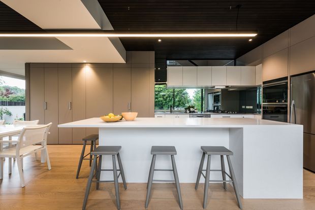 A spacious contemporary kitchen with a large white island and seating for four, integrated beige cabinetry, stainless-steel appliances, timber flooring, and a dark timber slatted ceiling. The open-plan space includes a dining area and extensive glazing overlooking greenery.