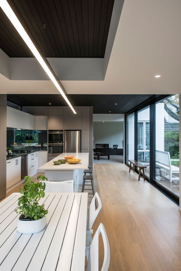 A bright, open-plan contemporary kitchen and dining area with a long white slatted dining table in the foreground, a white island with stools, and streamlined grey cabinetry with integrated stainless-steel appliances. The kitchen opens into a living room beyond and is bordered by extensive floor-to-ceiling glazing overlooking a garden. Dark timber-look slatted ceilings, pale timber flooring, crisp white walls, and linear recessed lighting create a refined, modern interior.