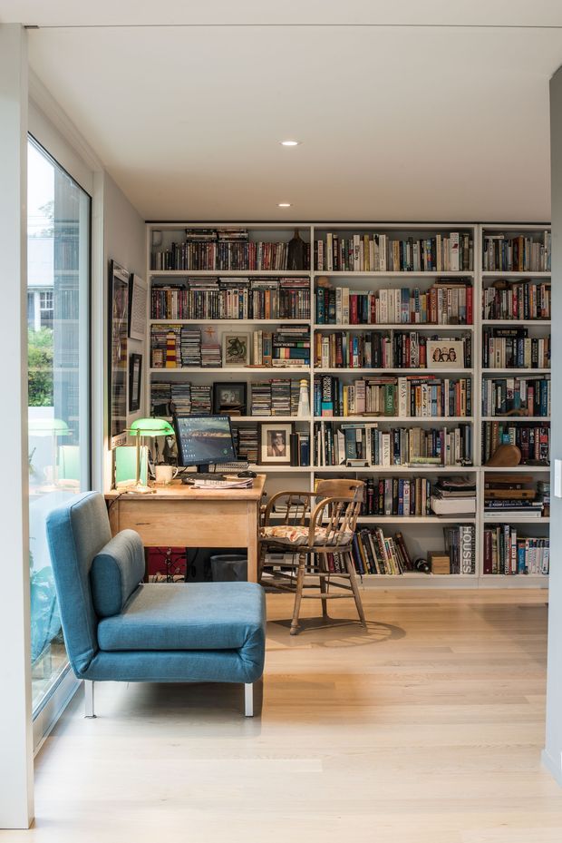 A bright, compact home office or study with a full wall of built-in white shelving filled with books, a timber desk and computer workspace, a wooden chair, and a blue upholstered armchair beside full-height glazing. The room has pale timber flooring, white walls and ceiling, recessed downlights, and a warm, book-lined atmosphere.