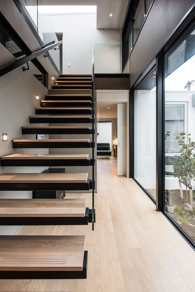 A contemporary interior hallway and stairway featuring a floating open-riser staircase with warm timber treads and dark metal supports. Full-height black-framed glazing runs along the right side, opening views to a small courtyard, while a double-height void, glass balustrade, recessed ceiling lights, and pale timber flooring create a bright, vertically expansive space. A living area is visible beyond the corridor.