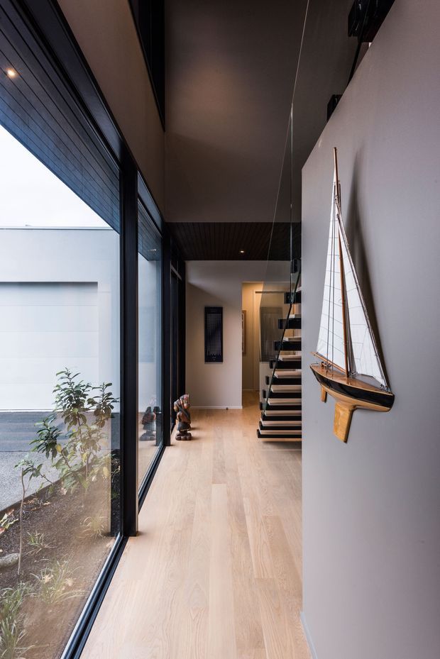 A narrow, contemporary residential hallway with pale timber flooring, full-height black-framed glazing on one side, and a minimalist white wall displaying a decorative model sailboat. A floating open-riser staircase with dark treads leads toward the rear of the home. The corridor has a dark ceiling section with recessed lighting, clean white walls, and views to a small planted courtyard through the glazing.