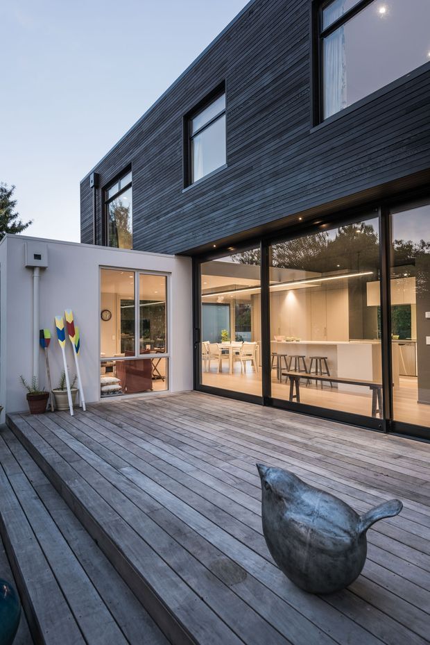 Exterior view of a contemporary two-storey home opening onto a large timber deck and courtyard. The dark horizontal cladding contrasts with pale rendered walls and extensive black-framed glazing, which reveals a warmly lit open-plan kitchen and dining area inside. The deck includes steps, outdoor seating, potted plants and decorative paddles.