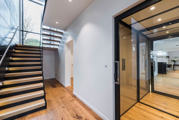 A bright contemporary entry hall with an open-riser timber staircase, black metal balustrade, expansive glazing, and warm timber flooring. A black-framed glazed internal door opens toward a kitchen or living area visible beyond. White walls, recessed ceiling lights, and strong vertical and horizontal lines create an airy, modern feel.