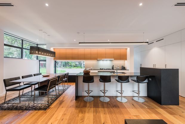 Open-plan contemporary kitchen and dining area with a long white island and five black bar stools, integrated white cabinetry, warm backlit timber cabinetry, stainless-steel appliances, and a large dining table beside expansive windows. The room has warm timber flooring, recessed lighting, and a linear pendant light over the dining table.