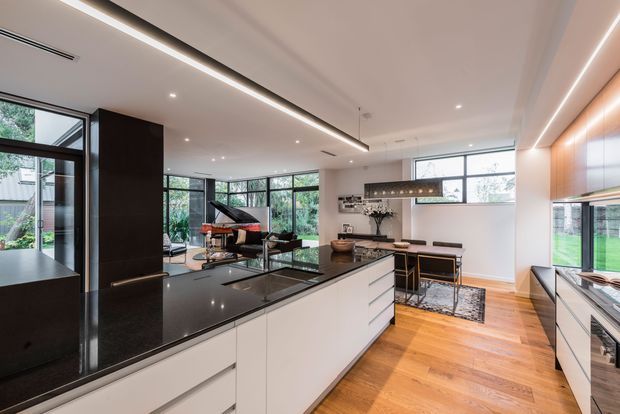 A sophisticated open-plan kitchen and dining area with a large white island, dark polished stone benchtops, and extensive black-framed glazing. Warm timber flooring contrasts with streamlined white cabinetry and dark feature joinery. The space includes a dining table, contemporary linear pendant lighting, and views into a living area with a grand piano.
