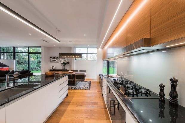 Contemporary open-plan kitchen with a long white island, dark stone benchtops, extensive timber overhead cabinetry, integrated linear lighting, and a large multi-burner gas cooktop. The kitchen opens to a dining and living area with garden views through large windows and glazed doors. Warm timber flooring contrasts with the white cabinetry and dark benchtops.