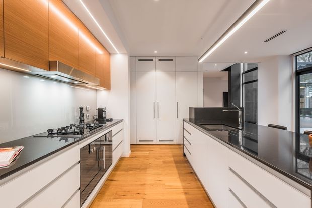 A contemporary galley-style kitchen with white handleless lower cabinetry, warm timber overhead cabinets, dark stone benchtops, integrated stainless-steel appliances, and a central island with seating. The room features linear recessed and under-cabinet lighting, timber flooring, a full-height white pantry or storage wall, and black-framed glazing along one side.