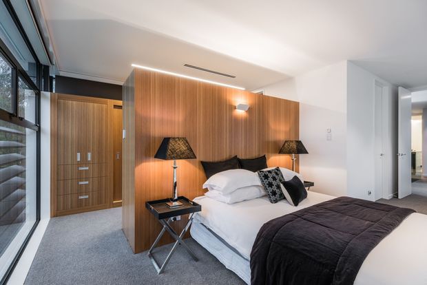 A contemporary bedroom with a queen-size bed dressed in white linens and a dark charcoal quilt, positioned against a warm vertical timber-panel feature wall. Matching bedside lamps, a dark bedside table, built-in timber wardrobe cabinetry, grey carpet, white walls, recessed ceiling ventilation, and large black-framed glazing create a refined, warm modern interior. An open doorway leads to an adjacent hall or room.