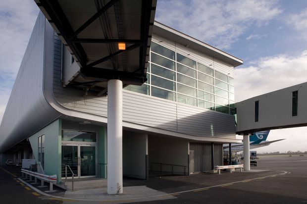 Exterior view of a modern airport terminal or commercial transport building, featuring a large glazed curtain-wall volume, pale horizontal metal cladding, sweeping curved facade elements, exposed structural steel canopies, and substantial cylindrical support columns. Aircraft and apron areas are visible beyond the building.