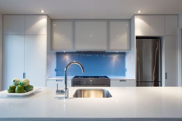 A bright, sleek contemporary kitchen viewed across a large white island benchtop. The kitchen features extensive white flat-panel cabinetry, a stainless-steel undermount sink with a tall curved faucet, a central range with stainless-steel hood area, a reflective blue horizontal-tile or glass splashback, and a large stainless-steel refrigerator integrated into the cabinetry. Recessed ceiling lighting and a clean uncluttered composition create a polished modern feel.