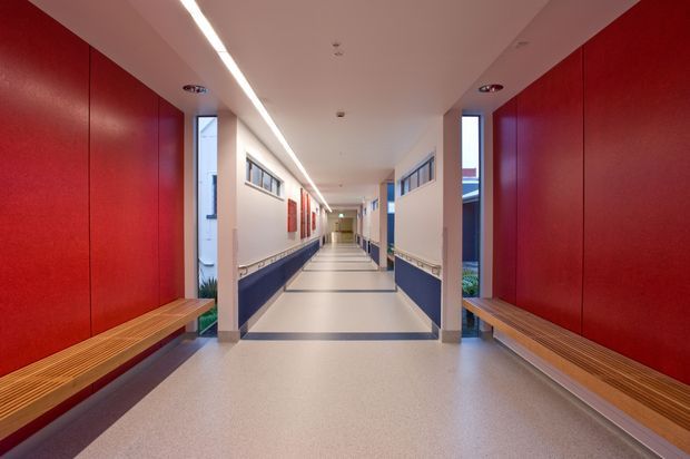 A long, bright institutional corridor with bold red wall panels, dark blue lower wall sections, pale flooring, narrow clerestory windows, continuous timber bench seating along both sides, and a linear recessed ceiling light. The space has a clean, modern, highly functional design with strong colour blocking and symmetrical perspective.