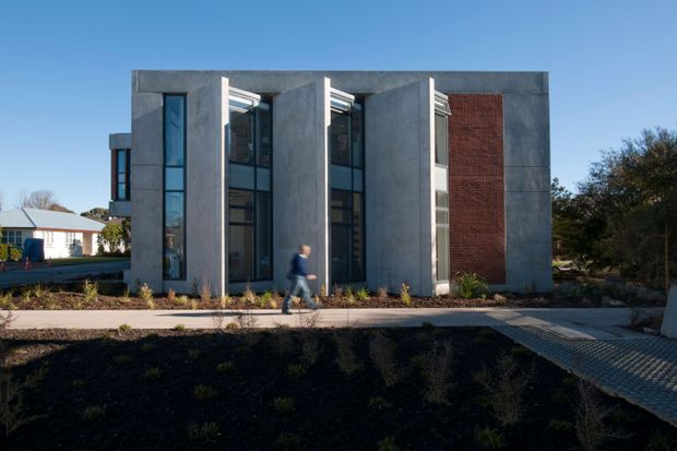Exterior view of a compact contemporary multi-storey building with a strongly rectilinear form. Tall, narrow glazed openings are recessed between projecting grey concrete-like wall planes, with a prominent red brick feature wall on the right. A paved pedestrian path and low landscaped planting sit in front, with a neighbouring low-rise building visible to the left.