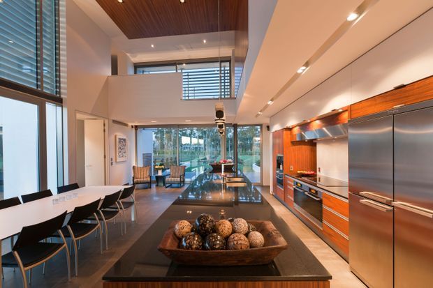 A spacious, high-end open-plan kitchen and dining area with a double-height volume and extensive glazing. The kitchen features warm timber cabinetry, stainless-steel appliances, a large dark island with a glossy surface, and integrated linear lighting. A dining table with black chairs sits to the left, while an upper-level opening and timber-lined ceiling add architectural interest. The room opens visually toward an outdoor garden or terrace through floor-to-ceiling glass.