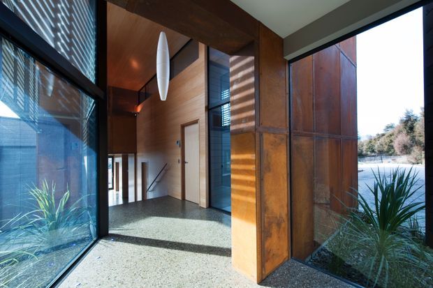 A contemporary entrance hall with expansive floor-to-ceiling glazing, warm timber wall and ceiling panels, rust-toned metal feature cladding, and a polished speckled floor. Indoor planting and views to landscaped water or garden areas are visible through the glazing. Strong linear forms, integrated blinds, and a sculptural pendant light create a refined, indoor-outdoor architectural character.