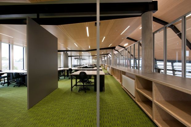 Open-plan contemporary office interior with rows of workstations, black-framed glazed partitions, exposed structural beams, timber-lined ceiling, green patterned carpet, and extensive built-in timber shelving and work surfaces. The space has abundant daylight and a long, efficient commercial layout.