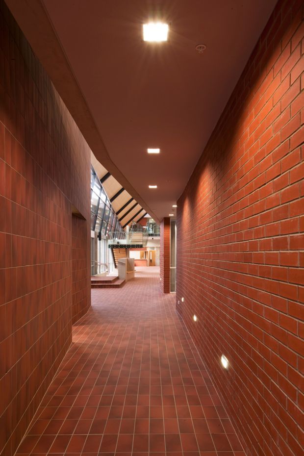A long, narrow interior corridor in a large institutional or commercial building, enclosed by red brick walls and dark red-brown tiled wall surfaces. Matching terracotta-red tiles cover the floor, while a smooth dark ceiling has recessed square downlights and small low-level wall lights. At the far end, the corridor opens into a brighter atrium with extensive glazing, exposed structural elements, steps, and a mezzanine walkway. The space has a strong brick-and-tile material palette and a warm, slightly dramatic atmosphere.