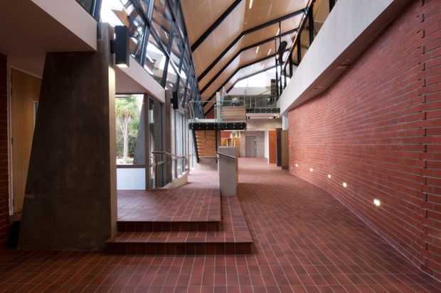 A spacious multi-level interior atrium or circulation hall with a dramatic glazed wall, exposed dark structural framing, warm timber-lined pitched ceiling, red brick feature wall, and extensive terracotta-red tiled flooring. Broad steps lead through the open space toward an upper-level mezzanine and bridge, with integrated wall lighting and planting visible beyond the glazing.