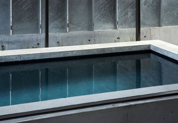 A close-up architectural view of a rectangular swimming pool enclosed by dark exposed concrete walls. The pool has clear blue water, a pale concrete coping, and strong geometric lines, creating a minimalist contemporary setting.