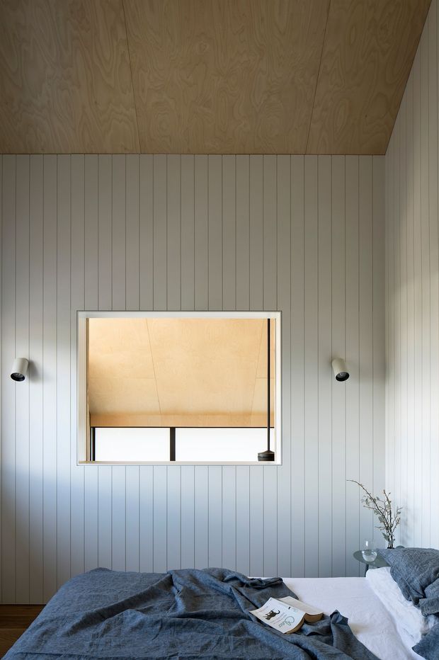 A calm, contemporary bedroom with full-height vertical wall panelling, a warm plywood-lined sloped ceiling, and a large framed interior window opening. The bed is dressed in muted blue-grey linen, with minimalist black-and-white wall sconces and a small bedside vase. The room has a restrained natural-material palette and clean built-in detailing.