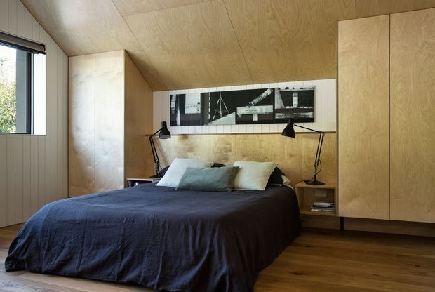A compact, warm bedroom lined extensively in light timber plywood, with built-in wardrobes and bedside storage, a low platform bed dressed in dark navy bedding, and a long horizontal black-and-white artwork above the headboard. Black articulated bedside lamps and a large window provide functional lighting and daylight. The space has a cohesive, minimalist timber interior with a sloping or angled ceiling plane.