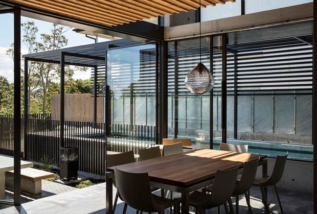 A contemporary indoor-outdoor dining area enclosed by extensive black-framed glazing. A large dark timber dining table with upholstered chairs sits beneath a striking timber slatted pergola ceiling, with a sculptural pendant light overhead. Beyond the glazing is a narrow water feature or pool, while vertical screening, concrete surfaces, and mature landscaping create a private courtyard atmosphere.