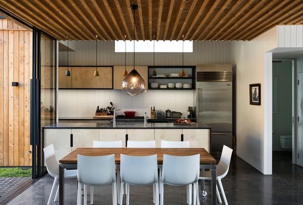 Contemporary open-plan kitchen and dining area with a long timber dining table, white dining chairs, a central island, stainless-steel appliances, and pale wood cabinetry. Full-height glazing and timber wall cladding appear on the left, while a richly detailed exposed timber slatted ceiling spans the room. The space has a clean, refined material palette with strong indoor-outdoor connection.